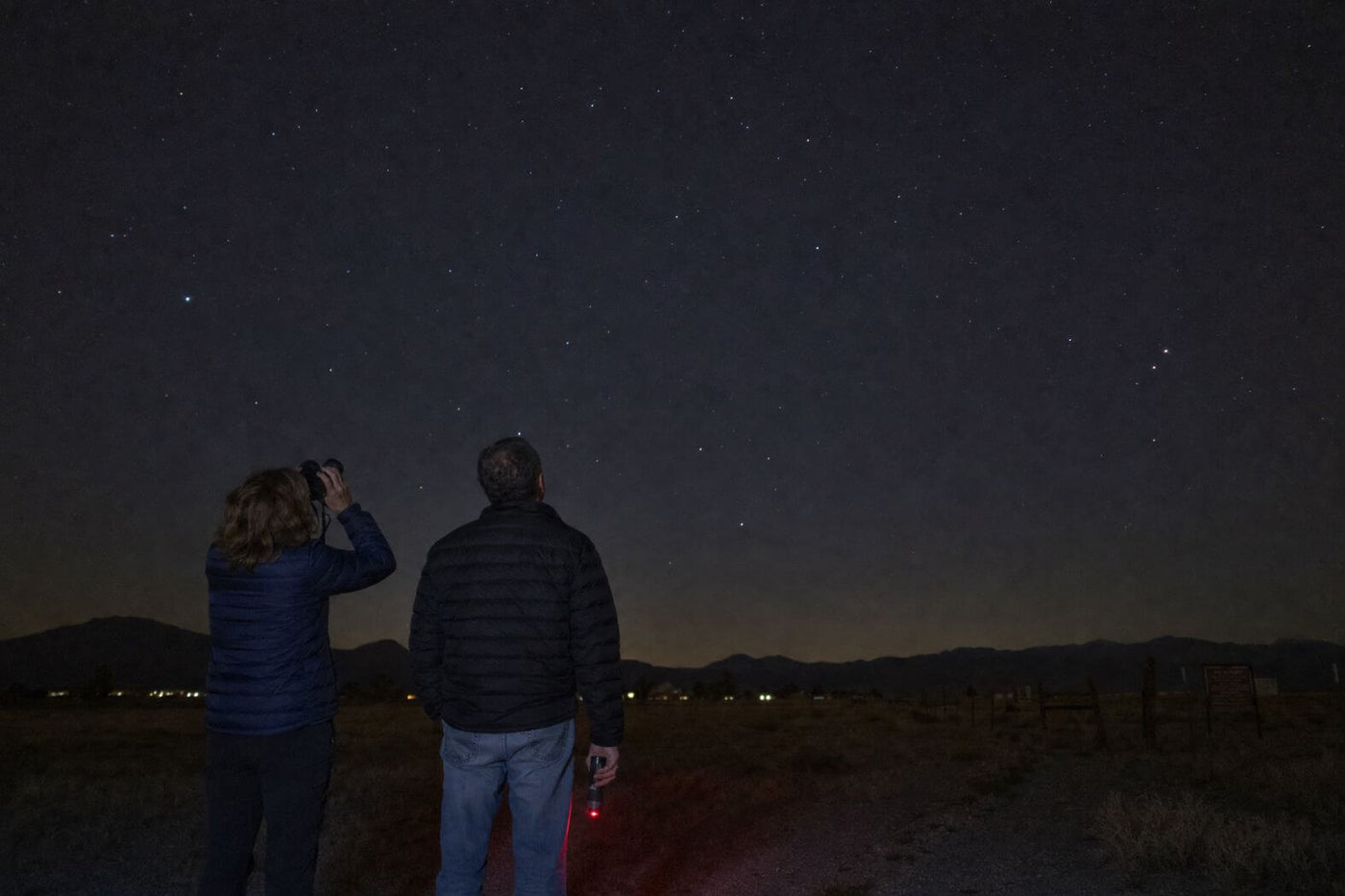 Two people observing stars with binoculars from a dark rural location